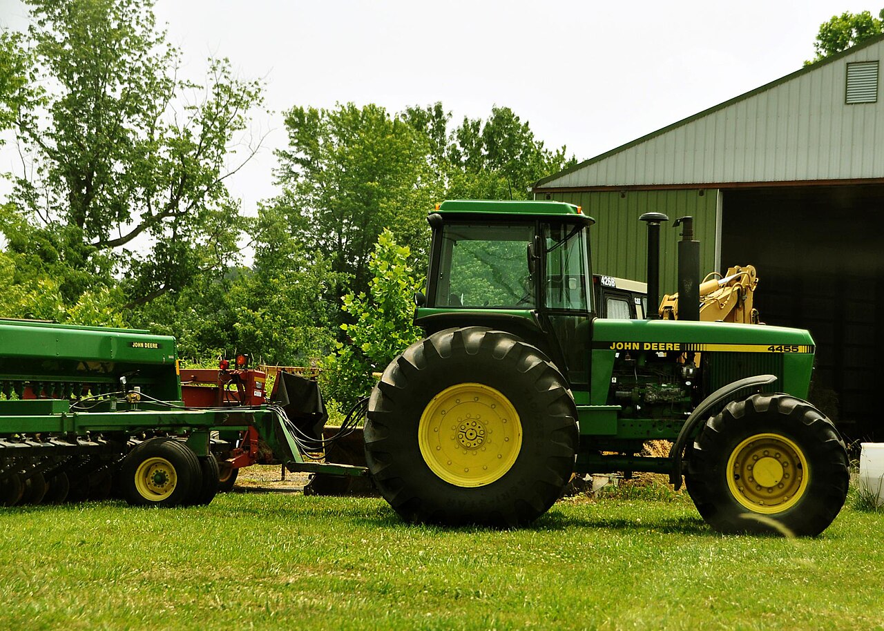 John Deere 4455 med John Deere-såmaskin fotograferad vid en gårdsbyggnad.