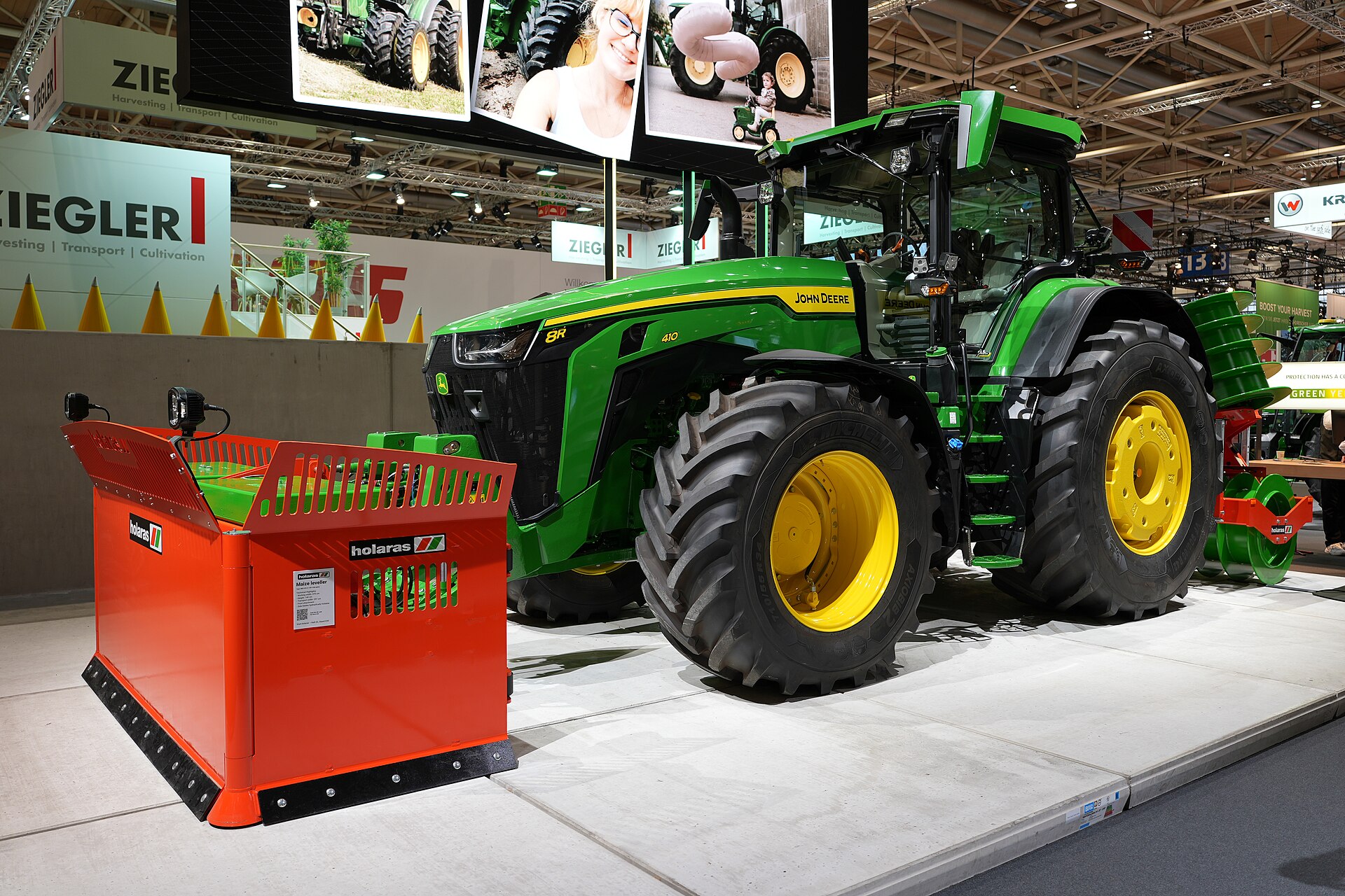 John Deere 8R 410 på mässa, med en frontmonterad skrapa och en vält baktill.