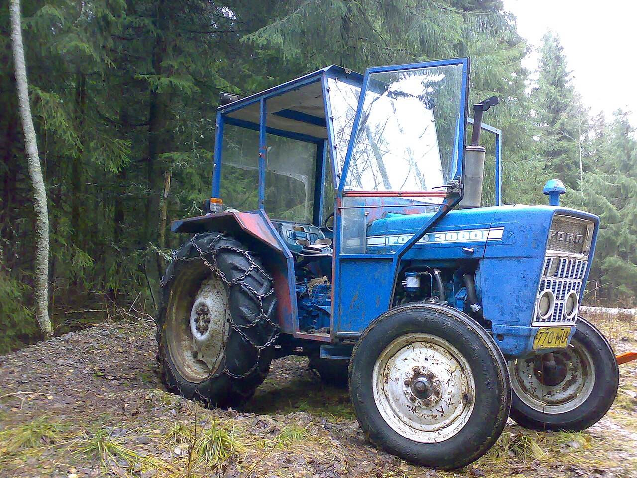 Blå Ford 3000-traktor sedd snett framifrån i skogskant; 3000-märket syns på motorhuven och kedjor sitter på bakhjulen.