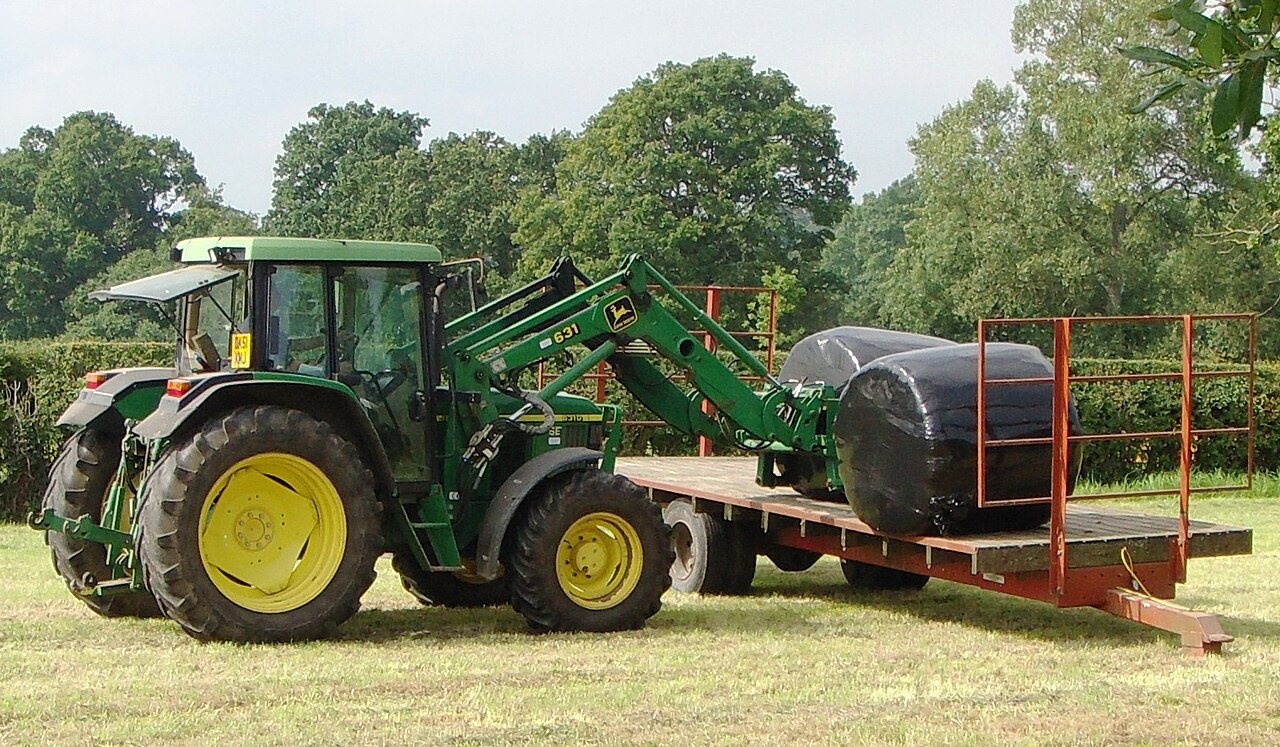 Grön John Deere 6310 SE med 631-frontlastare bredvid ett släp med ensilagebal i Wales.