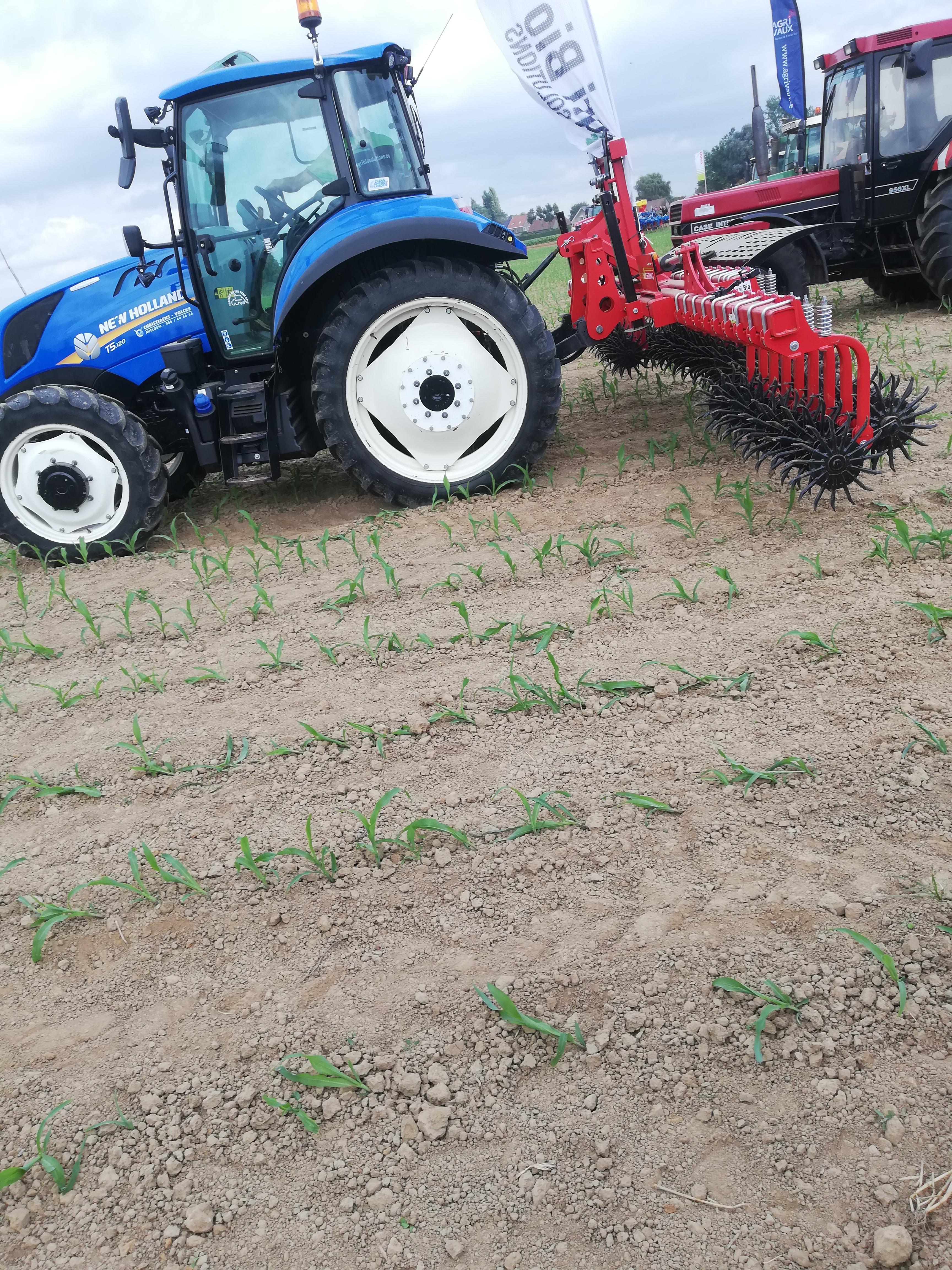 New Holland T5.120 med radhacka vid en maskinutställning; växellåda och generation framgår inte.