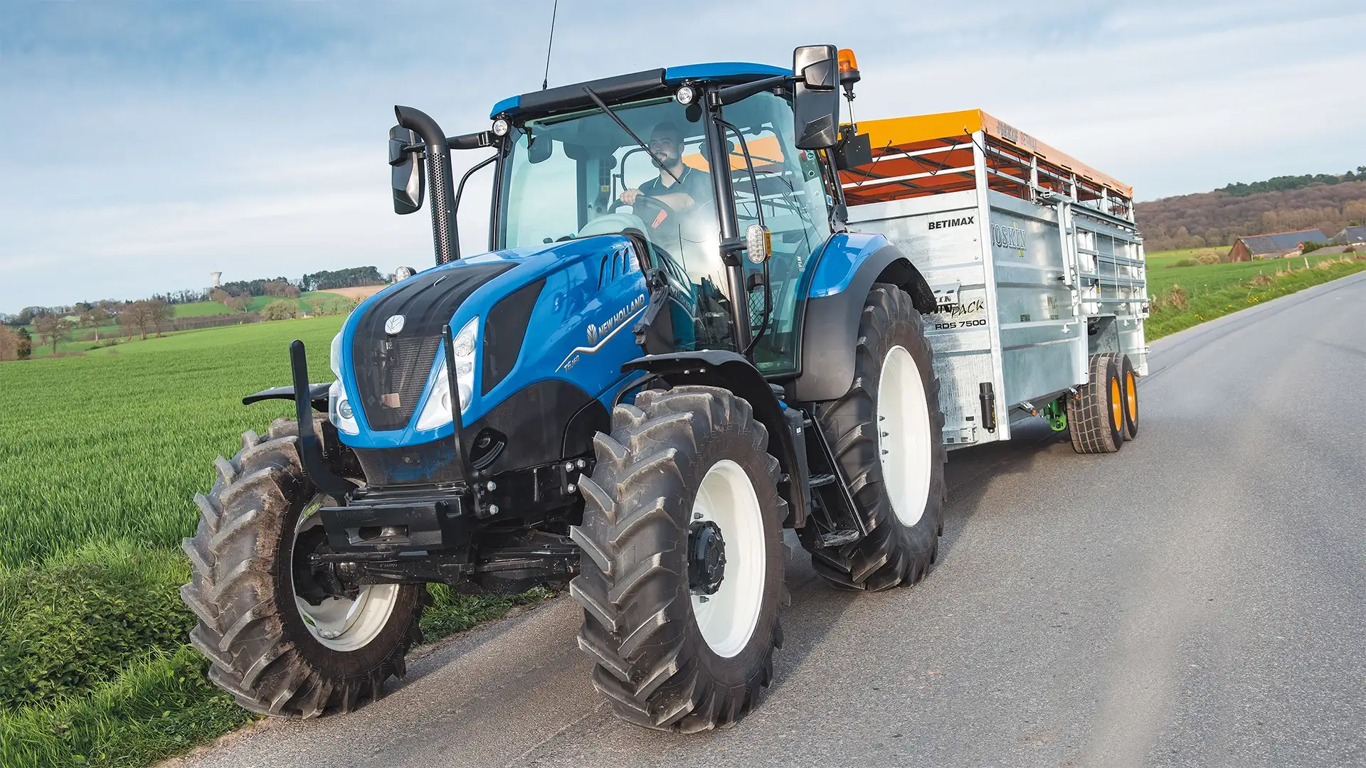 New Holland T6.160 med släp på landsväg; modellbeteckningen syns på motorhuven men transmissionen framgår inte.