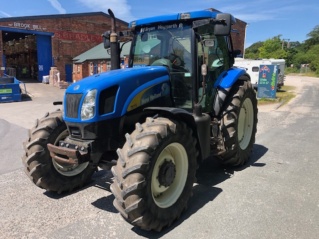 Blå New Holland TS115A-traktor fotograferad snett framifrån på en gårdsplan.